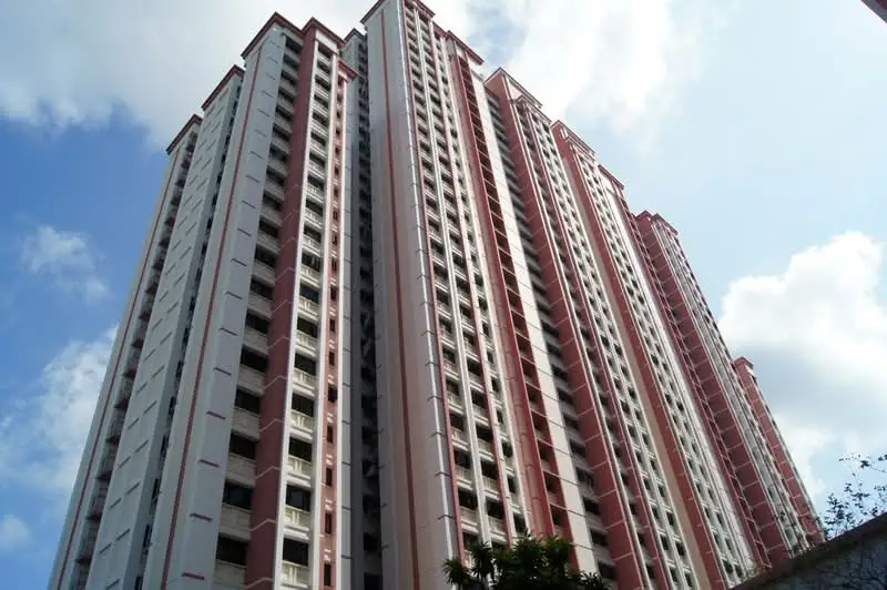 Low-angle view of a high-rise HDB apartment building complex in Singapore featuring red and off-white exterior facades under a partly cloudy blue sky.