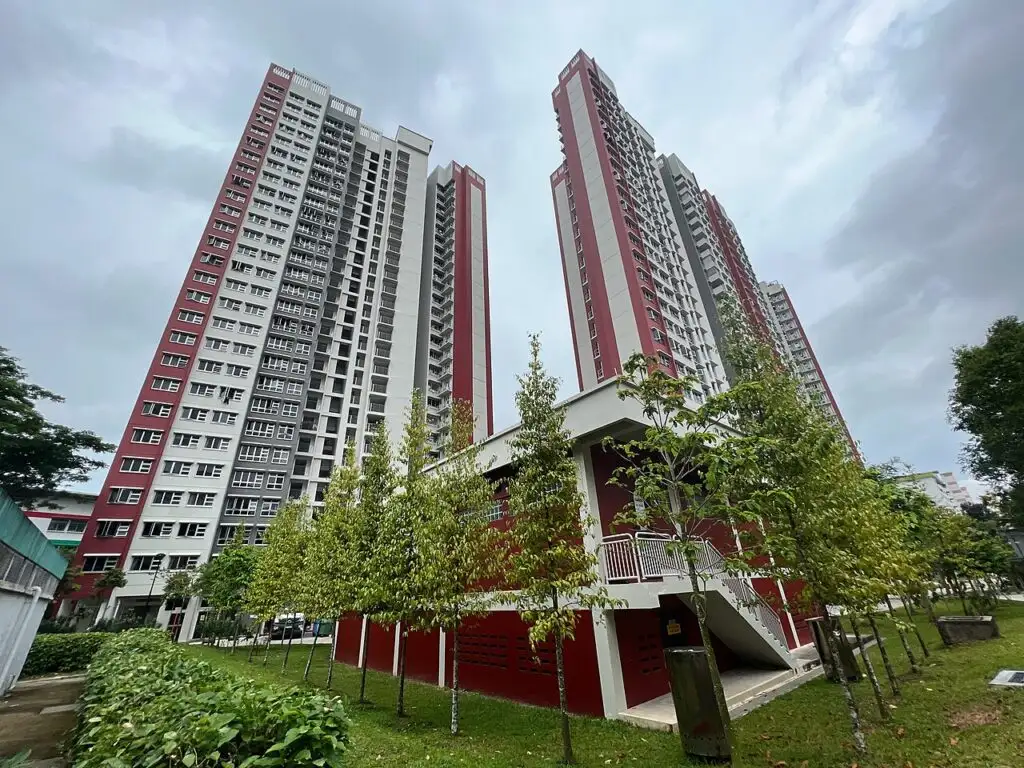 Exterior view of high-rise HDB apartment towers with a red, grey, and white color scheme, surrounded by lush green foliage and trees.