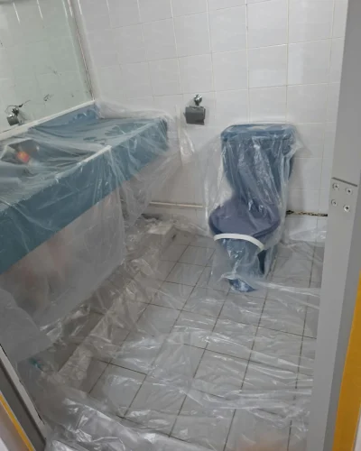 A bathroom with plastic sheet protection covering the toilet, vanity counter, and tiled floor in preparation for painting or renovation.