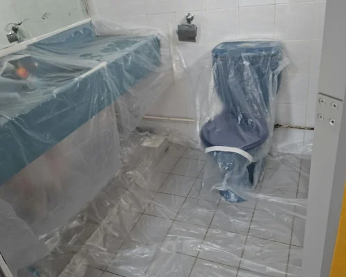 A bathroom with plastic sheet protection covering the toilet, vanity counter, and tiled floor in preparation for painting or renovation.