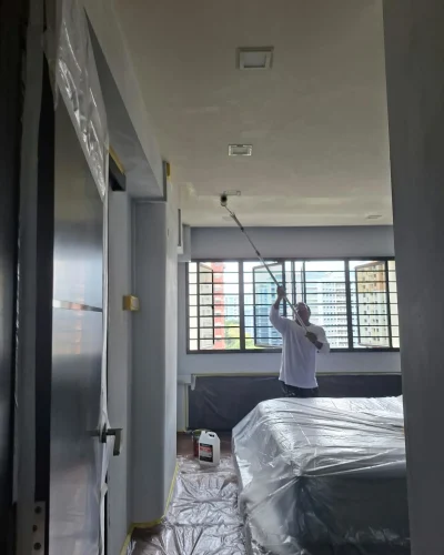 A painter in a long-sleeve white shirt using a roller on an extension pole to paint a bedroom ceiling white.