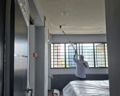 A painter in a long-sleeve white shirt using a roller on an extension pole to paint a bedroom ceiling white.