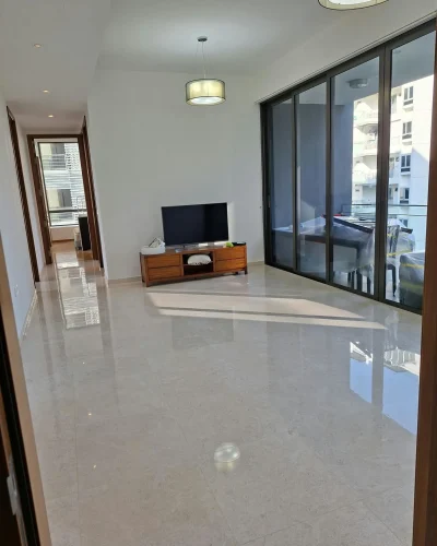 A spacious, brightly lit modern living room with polished marble floors, clean white walls, a TV media console, and large floor-to-ceiling glass sliding doors.