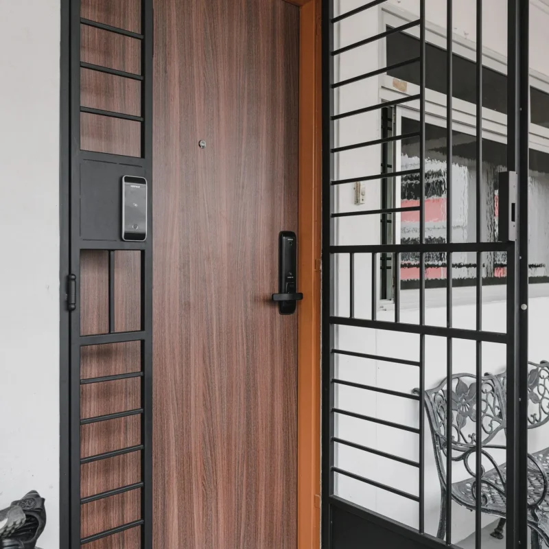 Modern HDB apartment main entrance featuring a wooden main door with a digital lock and a black metal wrought-iron security gate in Singapore.