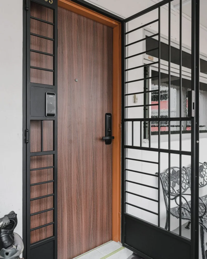 Modern HDB apartment main entrance featuring a wooden main door with a digital lock and a black metal wrought-iron security gate in Singapore.