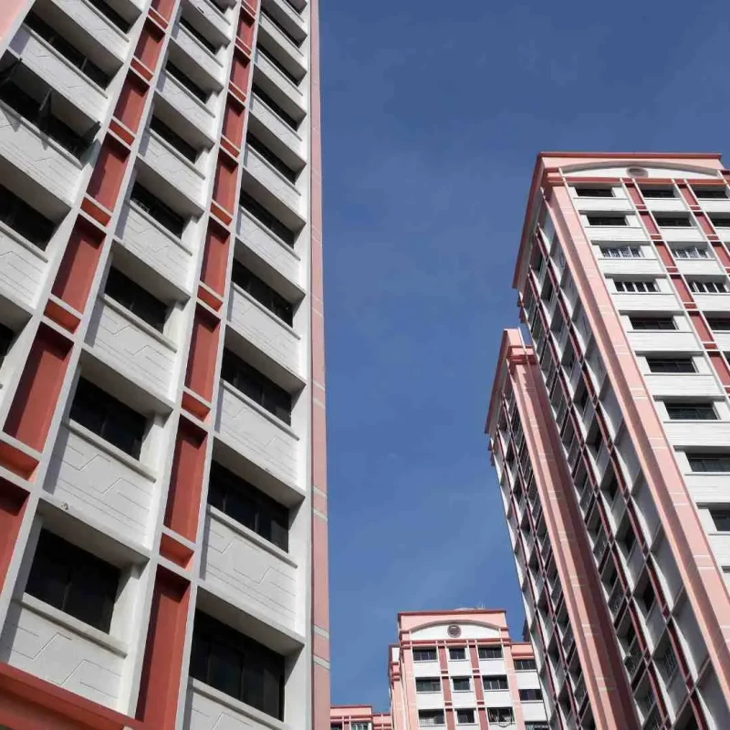 Low-angle shot looking up at tall HDB residential apartment towers painted in light pink, red accents, and white under a clear blue sky in Singapore.