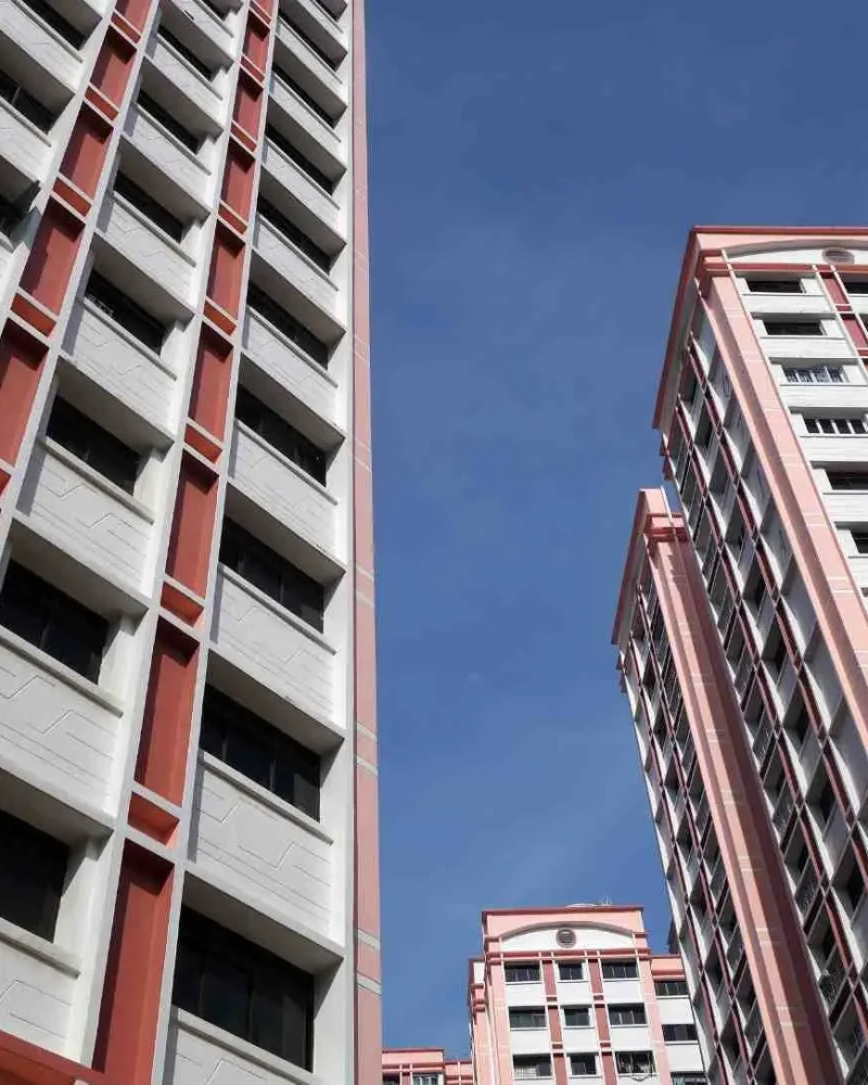 Low-angle shot looking up at tall HDB residential apartment towers painted in light pink, red accents, and white under a clear blue sky in Singapore.