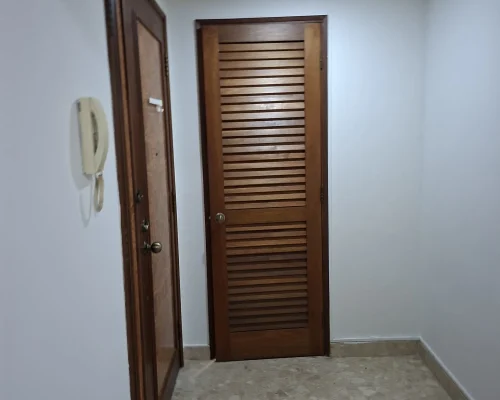 An interior hallway featuring smooth white walls, a wooden louvered door, a wooden frame door, and tiled flooring illuminated by a recessed ceiling light.