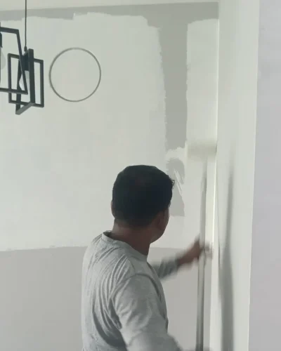 A painter in a long-sleeve grey shirt applying white paint to an interior wall with an extension roller.