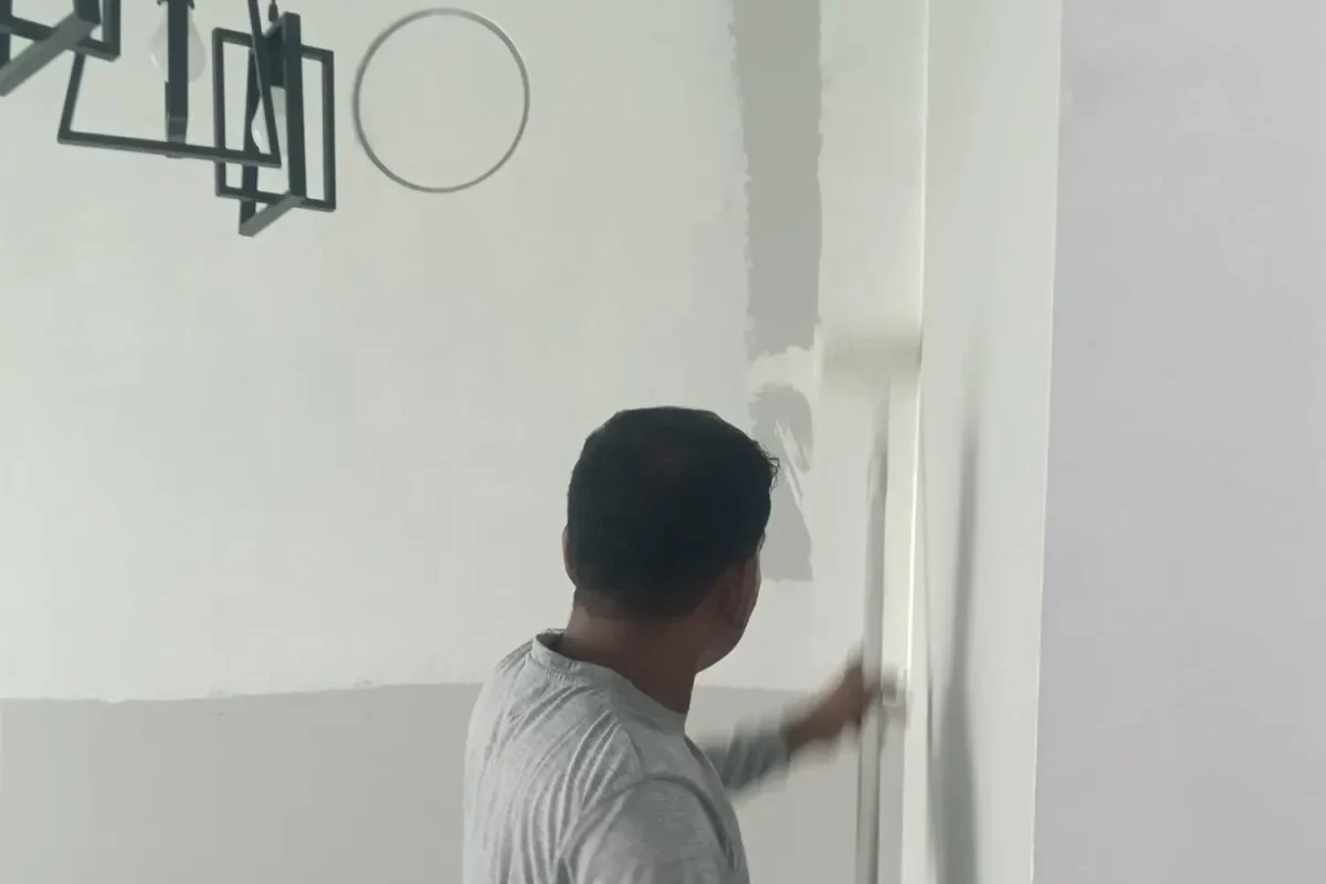 A painter in a long-sleeve grey shirt applying white paint to an interior wall with an extension roller.