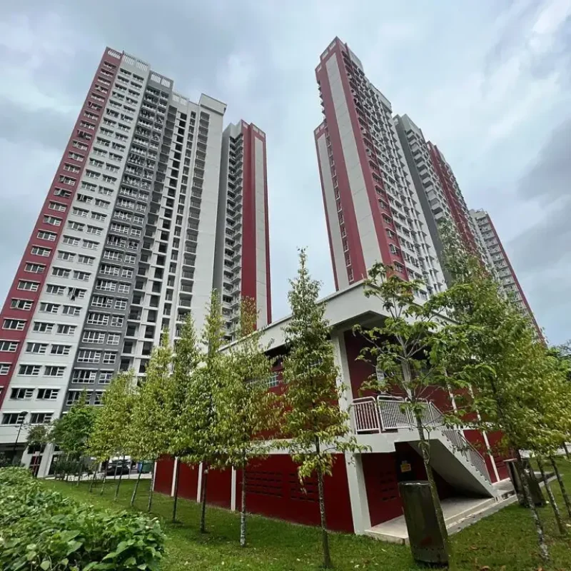 Exterior view of high-rise HDB apartment towers with a red, grey, and white color scheme, surrounded by lush green foliage and trees.